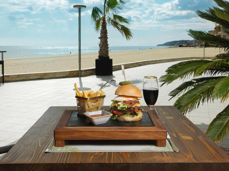 Tablett mit Burger, Pommes und Getränken am Strand mit Palme und Meer im Hintergrund.