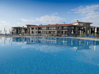 Großes Schwimmbecken mit klarem blauem Wasser, davor modernes Ferienhaus unter blauem Himmel.