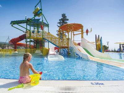 Wasserpark mit bunten Rutschen, Wasserattraktionen und einem Kind mit Schwimmflügel am Beckenrand.