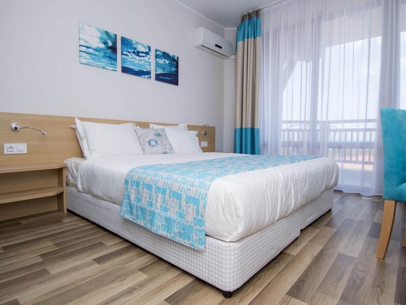 Helles Schlafzimmer mit Doppelbett, Holzfußboden, blauen Akzenten und großem Fenster mit Vorhängen.