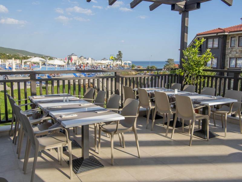 Terrasse mit mehreren Tischen und grauen Stühlen, Holzgeländer, Blick auf Gebäude und Meer.