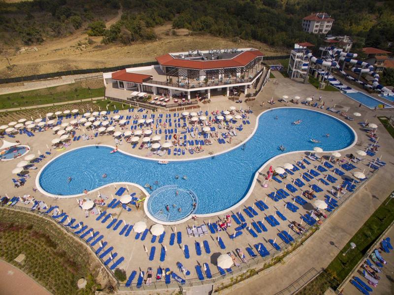 Großer, geschwungener Außenpool mit vielen blauen Liegestühlen und einem umliegenden Gebäude mit rotem Dach.