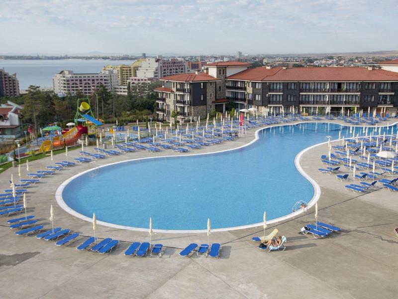 Großer, geschwungener Pool mit blauen Liegen und Schirmen vor Hotelgebäuden und Meer im Hintergrund.