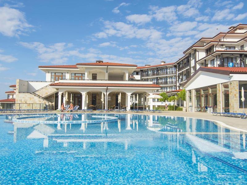 Großes Hotel mit blauen Pool, Sonnenschirmen und klarem Himmel im Hintergrund.