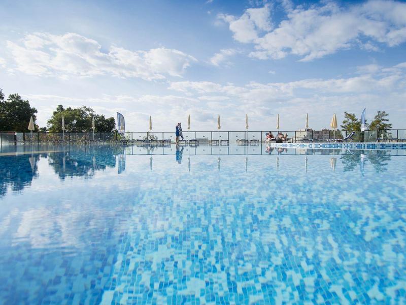 Ein klarer Pool mit blauer Mosaikfliese spiegelt den bewölkten Himmel und Bäume am Rand wider.