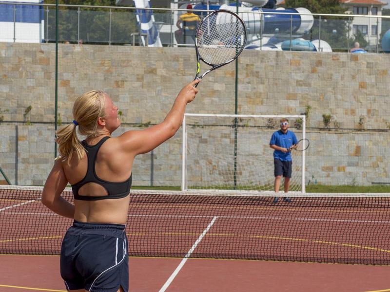 Frau in Sportkleidung spielt Tennis auf rotem Sandplatz bei sonnigem Wetter.