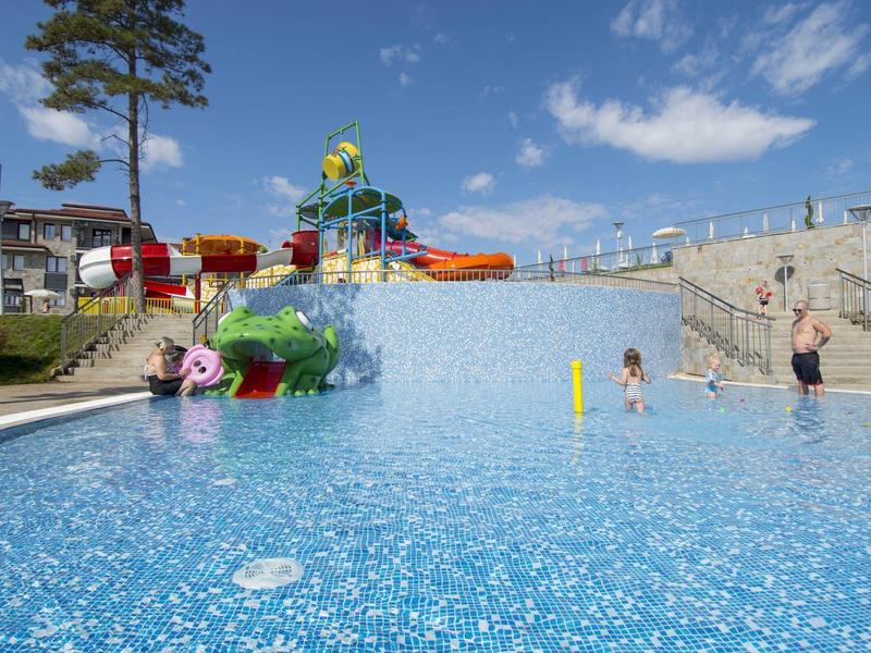 Kinder planschen im flachen Becken eines Outdoor-Wasserparks mit Rutschen und blauen Fliesen.