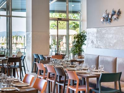 Modernes Restaurant mit Holztischen, verschiedenen Stühlen, großen Fenstern und dezentem Wanddekor.