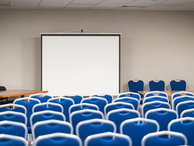 Sala conferenze vuota con sedie blu e schermo di proiezione bianco sulla parete.