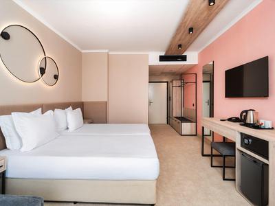 Camera d'hotel moderna con letto matrimoniale, illuminazione da parete e TV, arredata con colori caldi.