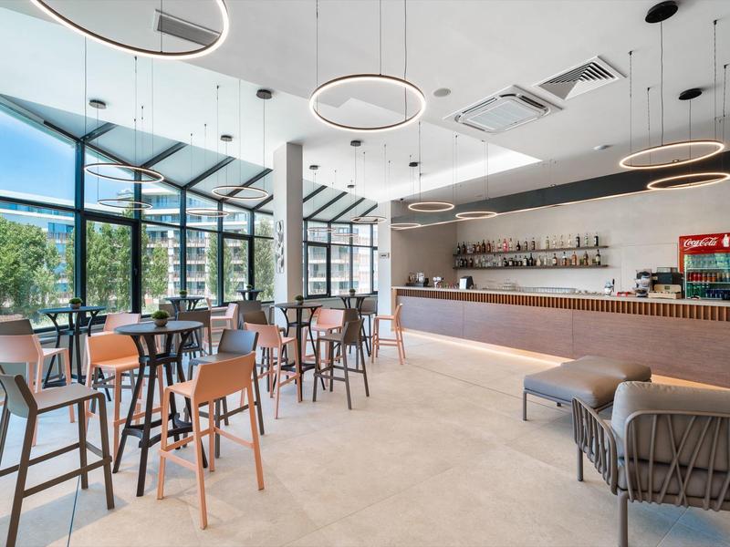 Caffè luminoso e moderno con grandi finestre, tavoli alti e area lounge.