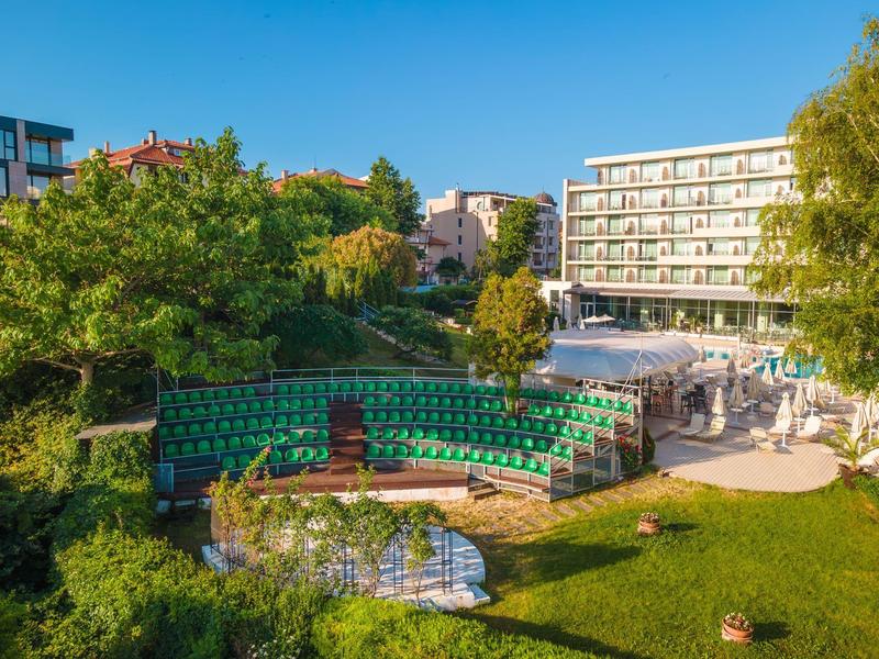 Complesso alberghiero moderno con giardino verde e palco all'aperto sotto un cielo limpido.