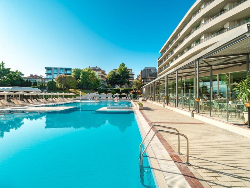Piscina moderna dell'hotel con lettini e facciata in vetro sotto un cielo sereno.