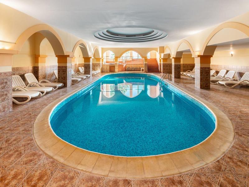 Area interna con piscina ovale, pavimenti marroni e lettini sotto archi curvi.