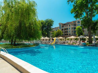 Großer blauer Hotelpool umgeben von Bäumen und Liegestühlen bei sonnigem Wetter.