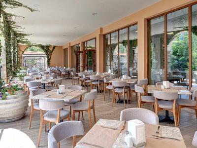 Überdachte Außenterrasse eines Hotels mit Tischen und Stühlen, Pflanzen und großen Fenstern.