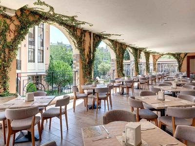 Ein heller, überdachter Außenbereich eines Restaurants mit Tischen, Stühlen und Grünpflanzen an den Säulen.