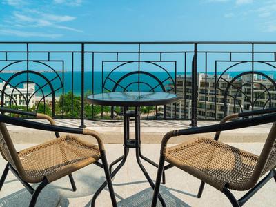 Balkon mit zwei Stühlen und kleinem Tisch mit Blick aufs Meer und Gebäude bei klarem Himmel.