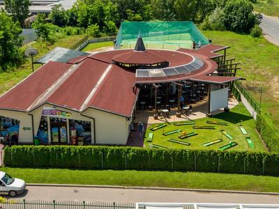 Kruhová budova s červenou střechou a venkovním minigolfem obklopená zelení a vozidly.