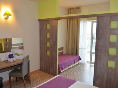 Moderne hotelkamer met paarse bedden, houten bureau, stoel en schuifwand naar balkon.