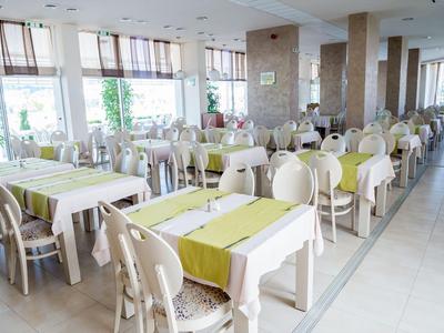 Světlá jídelna s mnoha bílými stoly a židlemi, zelené ubrusy a velká okna.