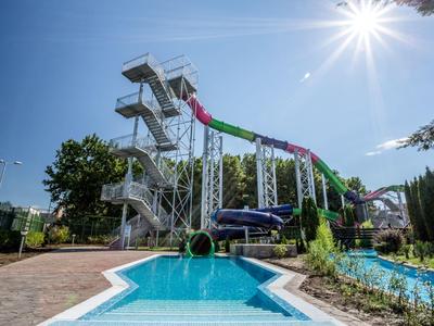 Barevná věž s tobogány a bazénem, obklopená stromy pod slunečnou oblohou.