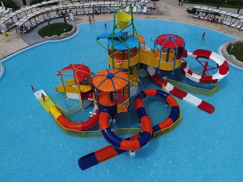 Grote buitenspeelstructuur met glijbanen en waterspuiten in een zwembad bij een resort.
