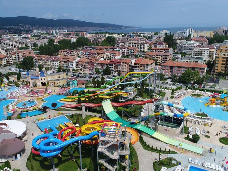 Groot waterpark met glijbanen en zwembaden bij stadsgebouwen en heuvels op de achtergrond.