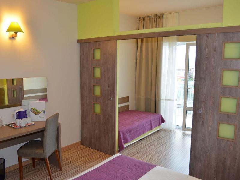 Moderne hotelkamer met paarse bedden, houten bureau, stoel en schuifwand naar balkon.