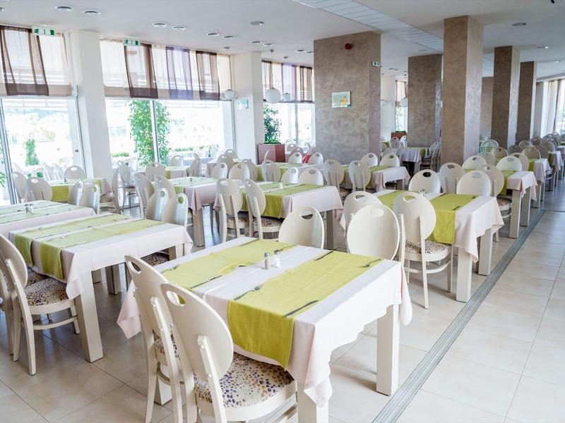 Lichte eetzaal met witte tafels en stoelen, groene tafellopers en grote ramen.