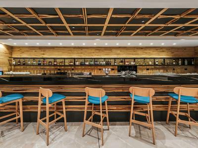 Espace bar d'hôtel moderne avec des tabourets en bois et des étagères éclairées remplies de bouteilles.