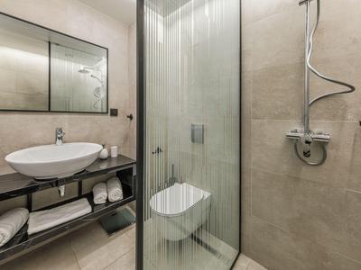 Salle de bain d'hôtel moderne avec douche vitrée, WC mural, lavabo sur étagère ouverte et serviettes en dessous.