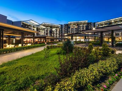 Modernes Hotelgebäude mit Beleuchtung, umgeben von gepflegtem Garten und klar blauem Abendhimmel.