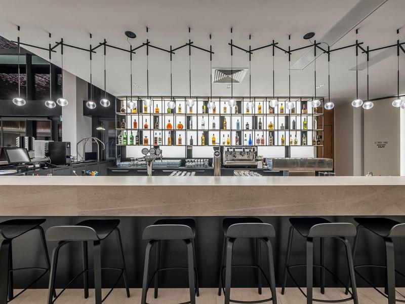 Bar moderne d'hôtel avec long comptoir en bois, tabourets noirs et étagère éclairée en arrière-plan.