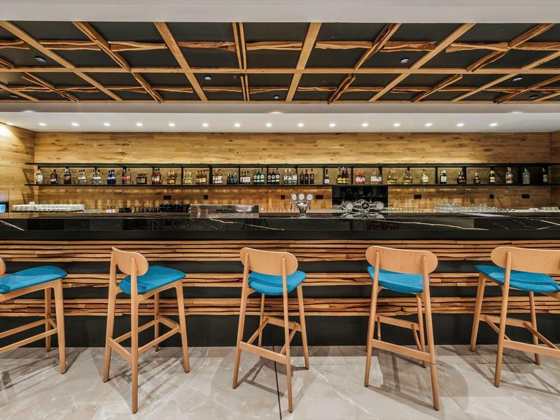 Espace bar d'hôtel moderne avec des tabourets en bois et des étagères éclairées remplies de bouteilles.