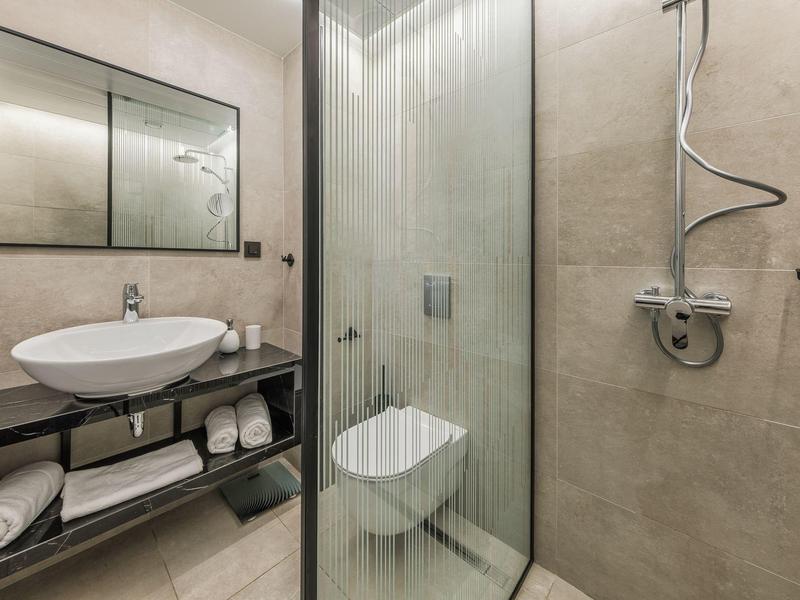 Salle de bain d'hôtel moderne avec douche vitrée, WC mural, lavabo sur étagère ouverte et serviettes en dessous.