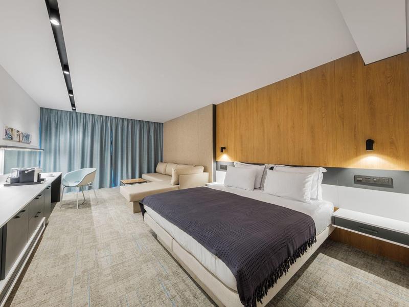 Chambre d'hôtel moderne avec lit double, mur d'accent en bois, canapé et rideaux gris.