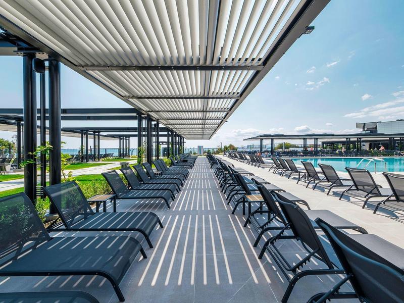 Espace piscine moderne avec rangées de chaises longues et protection solaire sous un ciel bleu.
