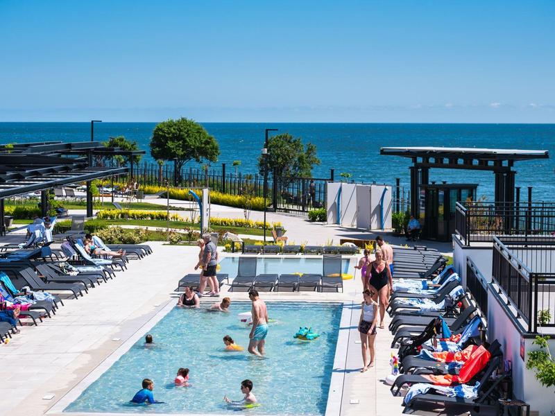 Zone de piscine avec des personnes, des chaises longues et vue sur la mer et le ciel bleu.