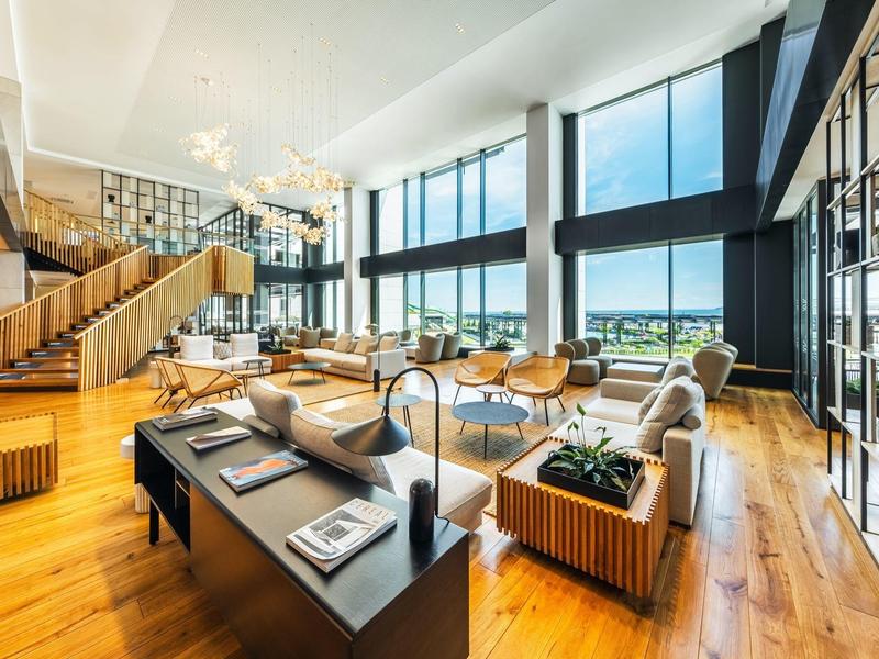 Grand salon d'hôtel lumineux avec fenêtres panoramiques et design en bois moderne.