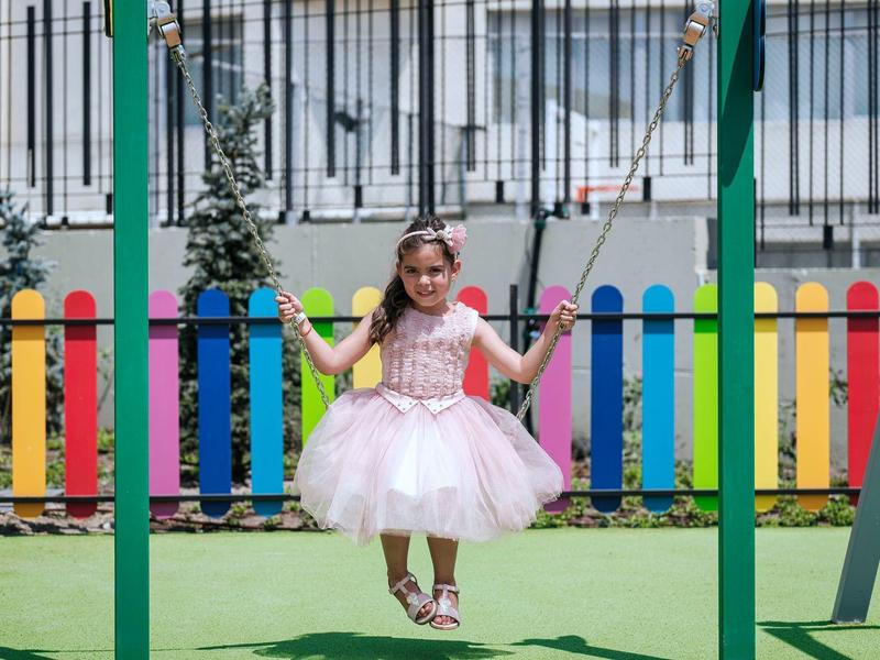 Petite fille en robe rose sur une balançoire dans une aire de jeux avec une clôture colorée.