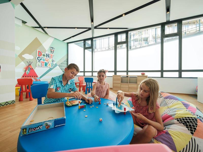 Des enfants jouent à une table bleue dans une salle de jeux moderne et lumineuse avec de grandes fenêtres.
