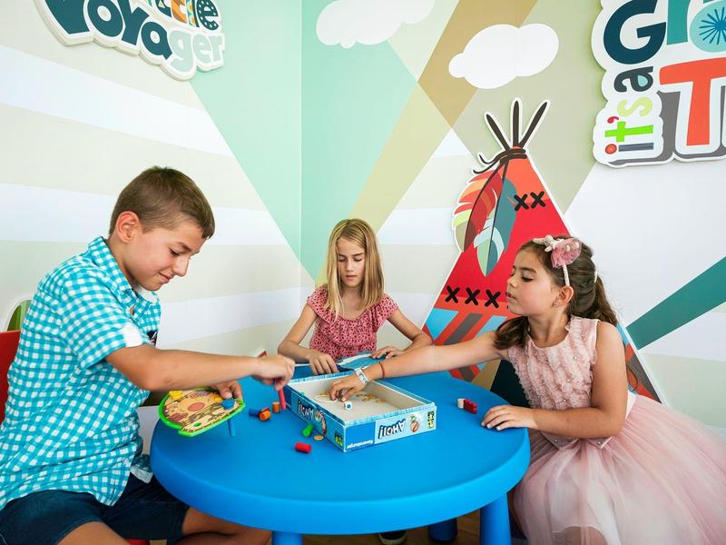 Trois enfants jouent à une table bleue dans une pièce colorée avec un tipi peint au mur.