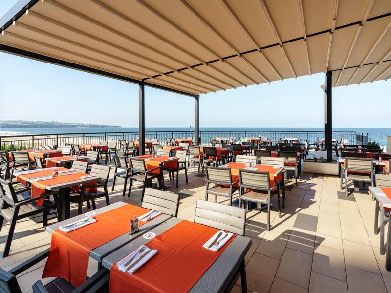 Open restaurant met oranje tafelkleden en zeezicht onder een luifel.