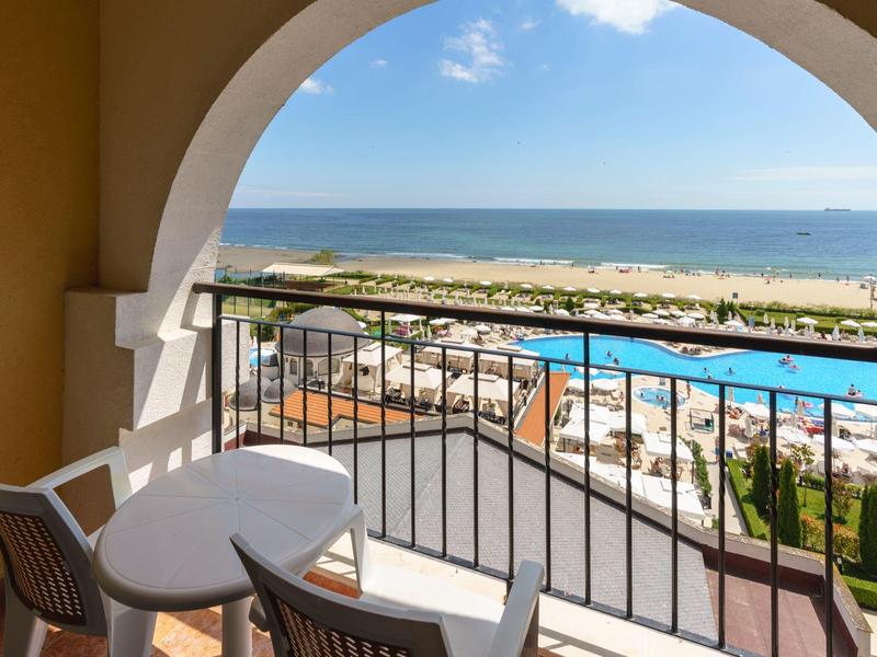 Balkon met tafel en stoelen, uitzicht op zwembad, strand en zee onder een heldere hemel.