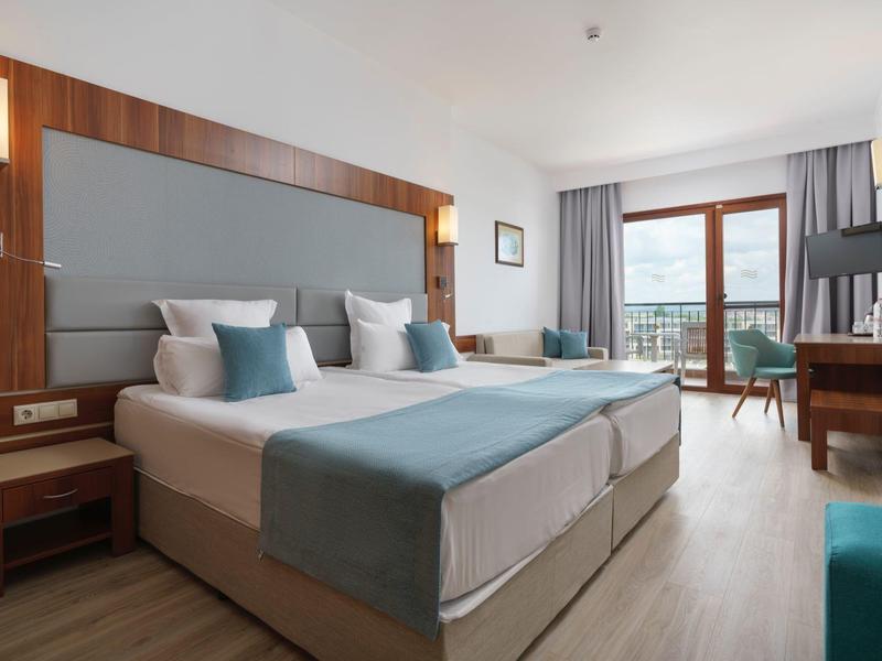 Lichte hotelkamer met twee bedden, houten meubels en balkon met uitzicht.