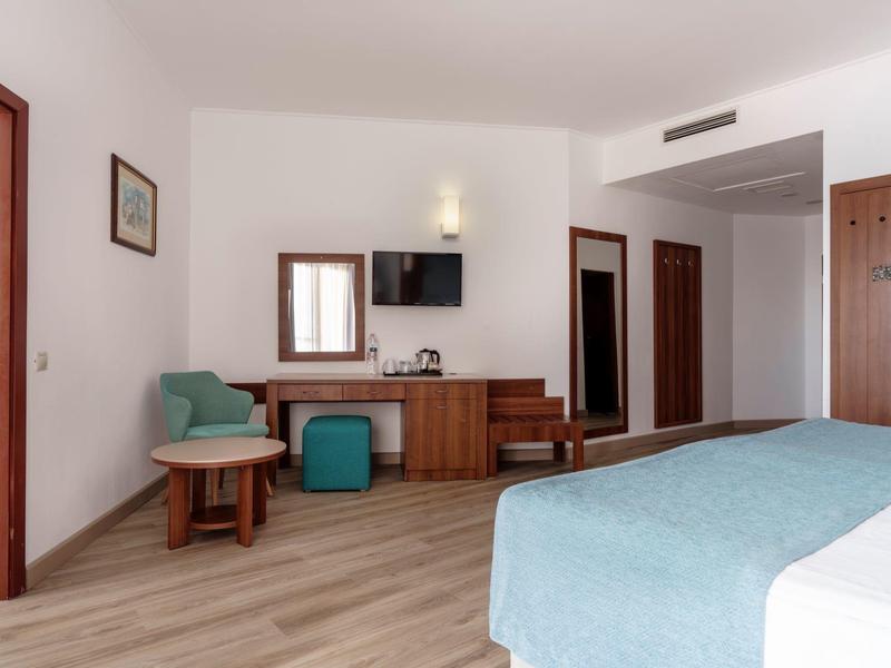 Lichte hotelkamer met houten vloer, blauw bed, groene fauteuil en aan de muur gemonteerde tv.