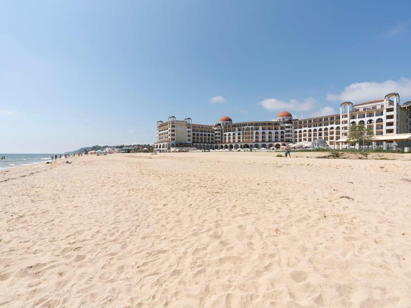 Breed zandstrand met hotelcomplex aan de horizon onder een blauwe lucht.