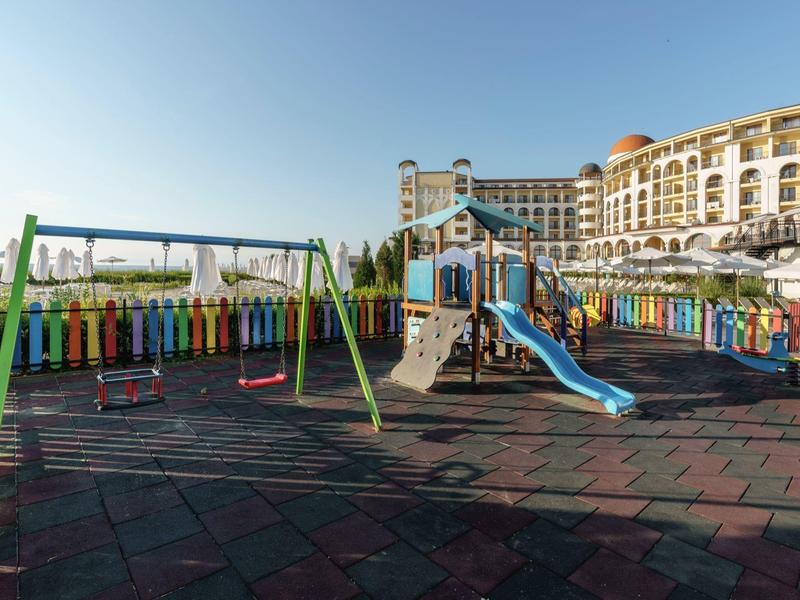 Kleurrijke kinderspeelplaats met schommels en glijbaan naast een groot hotel onder een heldere lucht.