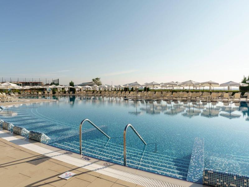 Groot rond hotelzwembad met helder blauw water en talrijke ligstoelen onder parasols.
